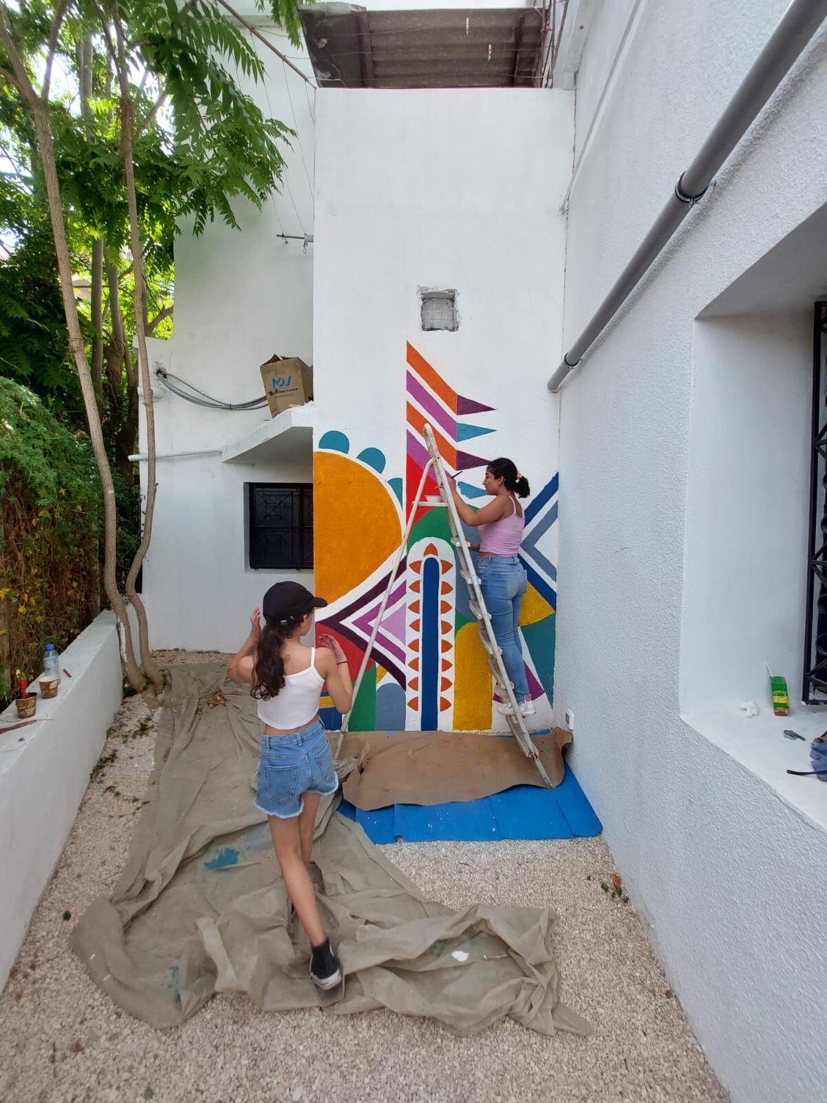 Two volunteers painting a large colorful geometric mural on an exterior building corner