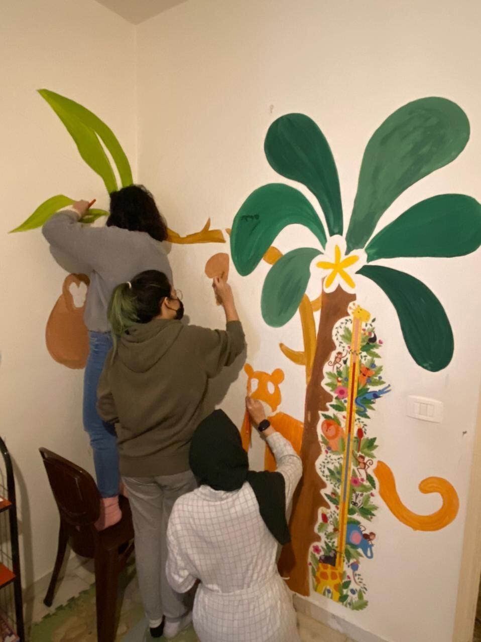 Volunteers painting a jungle-themed mural with palm trees and animals in a children's room