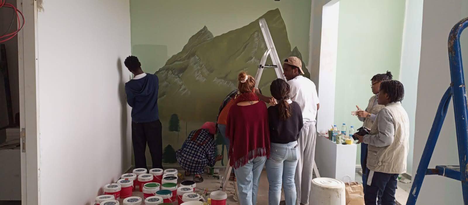 A group of volunteers gathered around paint cans, painting a mountain landscape mural together
