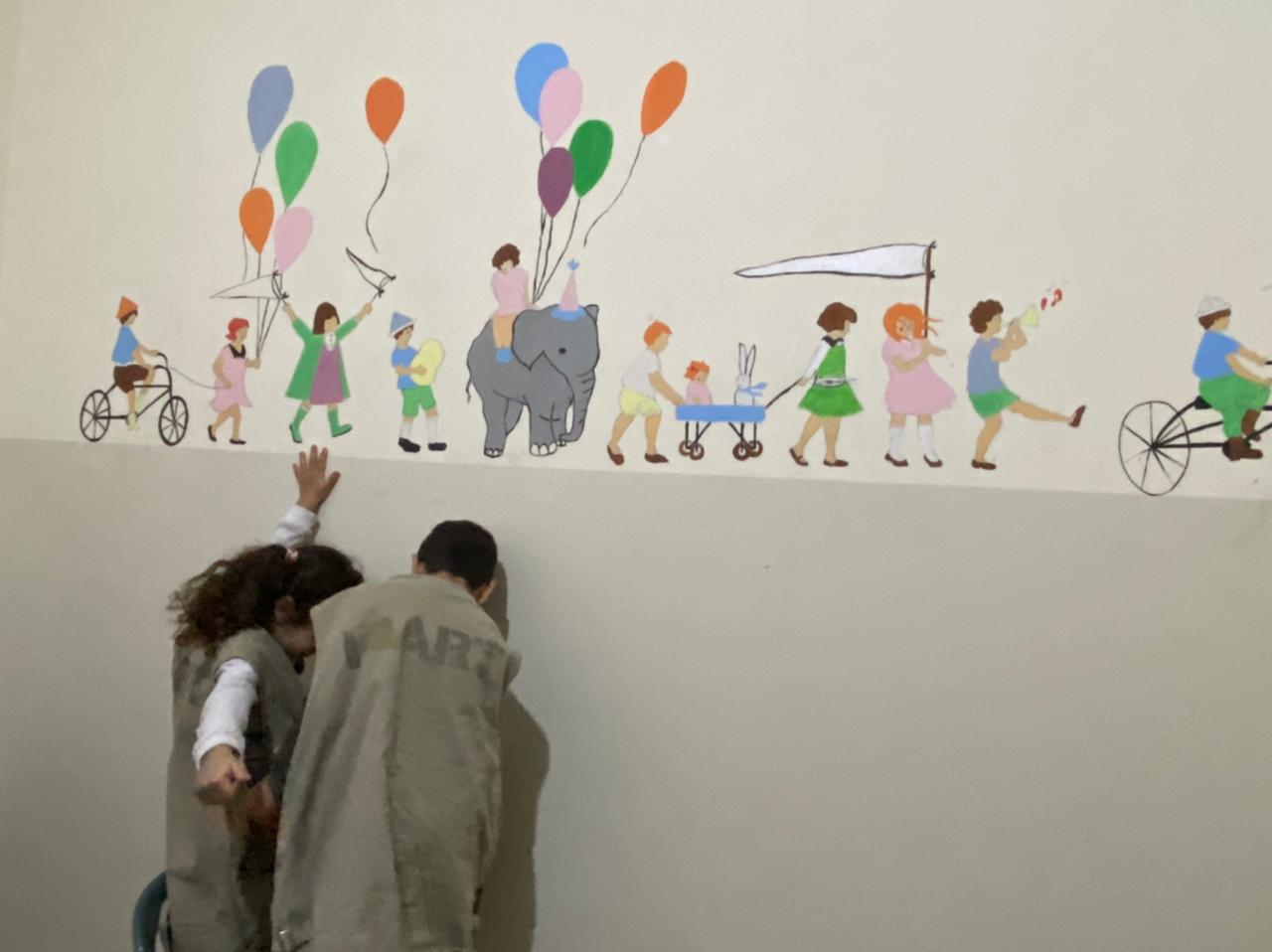 Two volunteers finishing a whimsical parade-of-children mural with balloons and an elephant