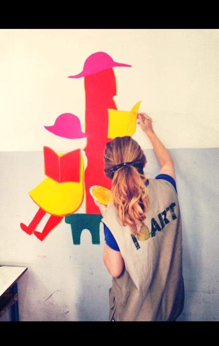 A volunteer in an I Leaf Art vest painting a reading-themed mural of a girl on a book