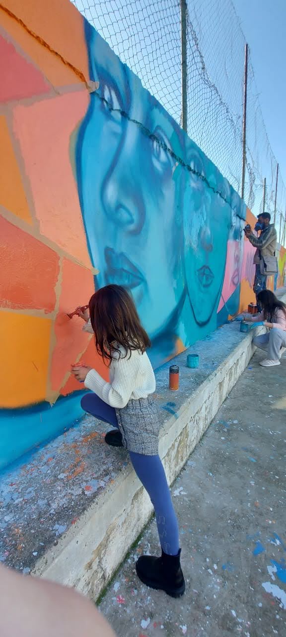 A volunteer painting a large-scale blue portrait mural on an outdoor fenced wall