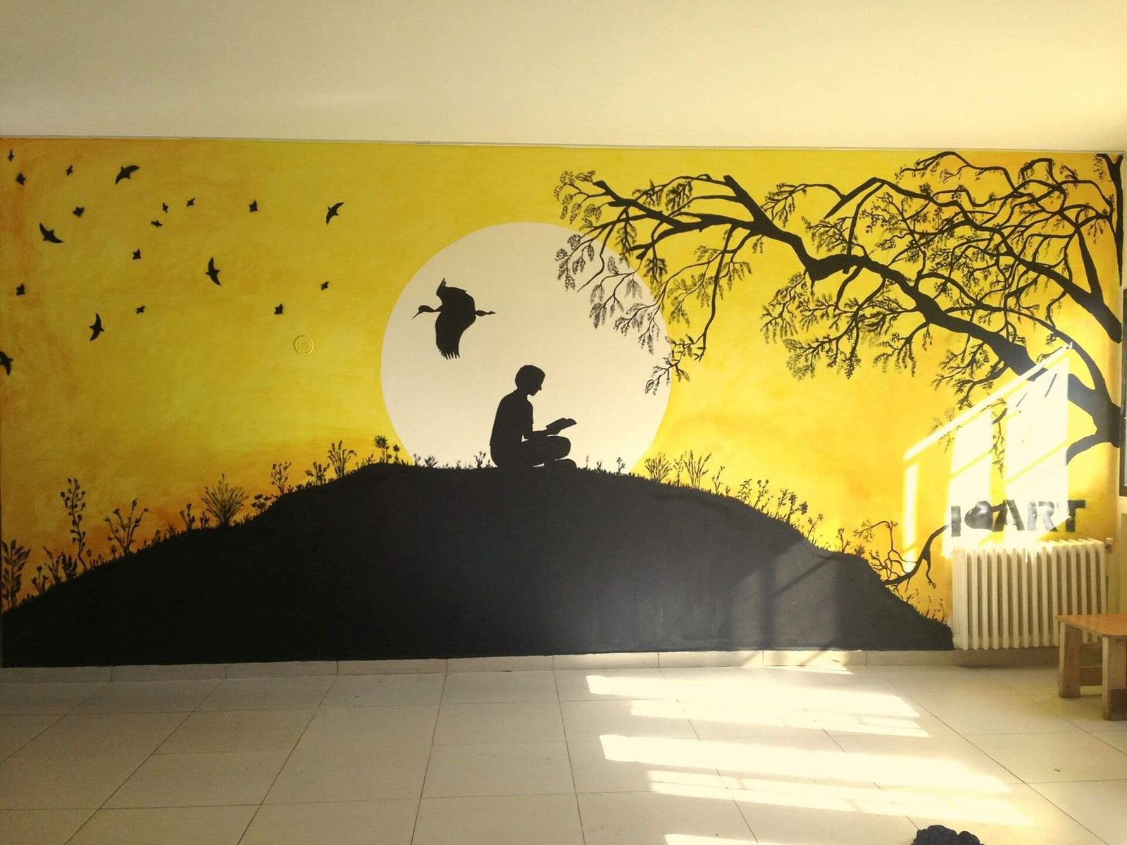 A silhouette mural of a person reading on a hill at sunset, with a bird flying overhead and a tree branch