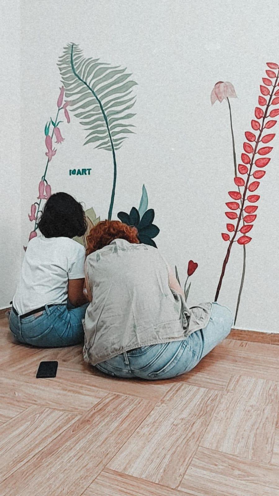 Two people sitting on the floor in front of a wildflower wall illustration